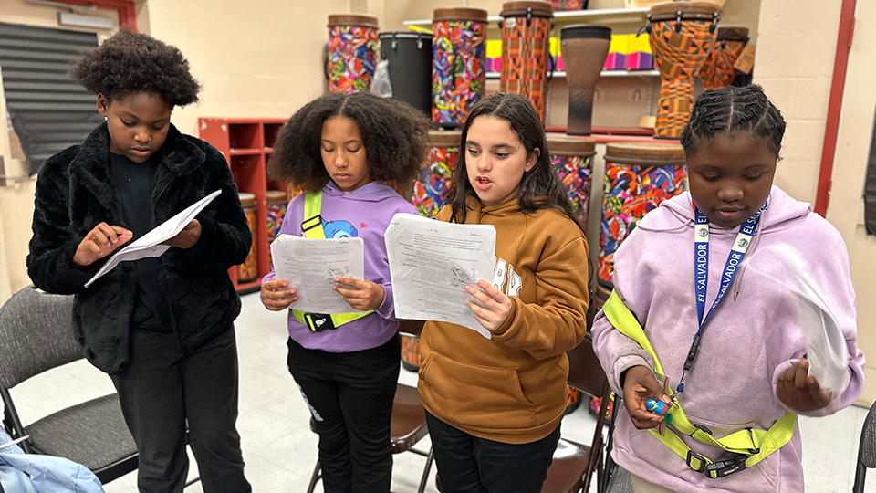 Four students standing in a classroom, reading theatre scripts