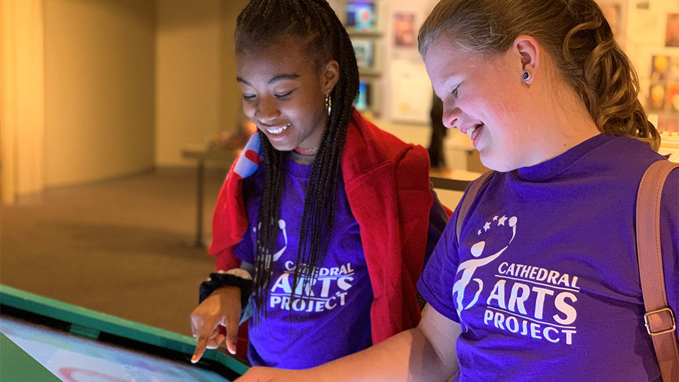 Two students in purple shirts interacting with a touch screen art display
