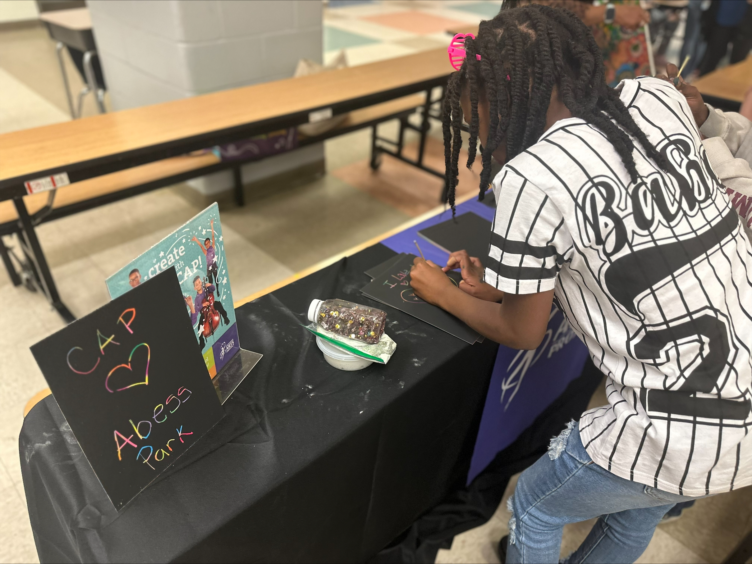 A student makes art on a table where a handmade sign reads "CAP hearts Abess Park"